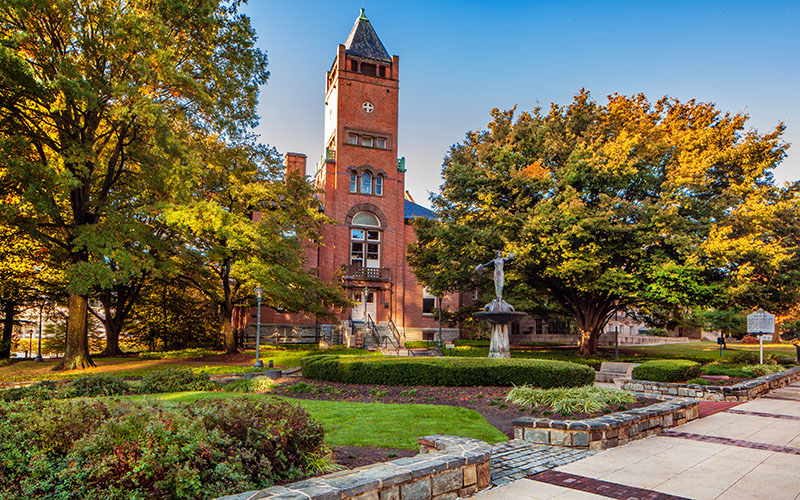 A red brick building with a clock tower is surrounded by green lawns, trees, and pathways under a clear blue sky.