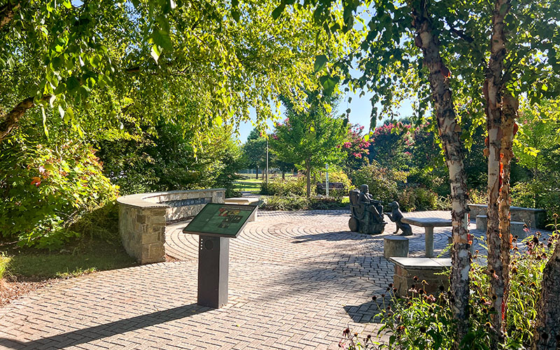 A sunlit park scene with a curved stone bench, an informational sign, and a bronze sculpture of people sitting on benches surrounded by trees and greenery.