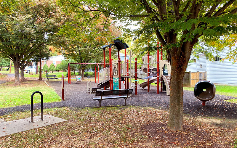 A playground with climbing structures, slides, and swings is surrounded by trees and benches, with a bike rack in the foreground.