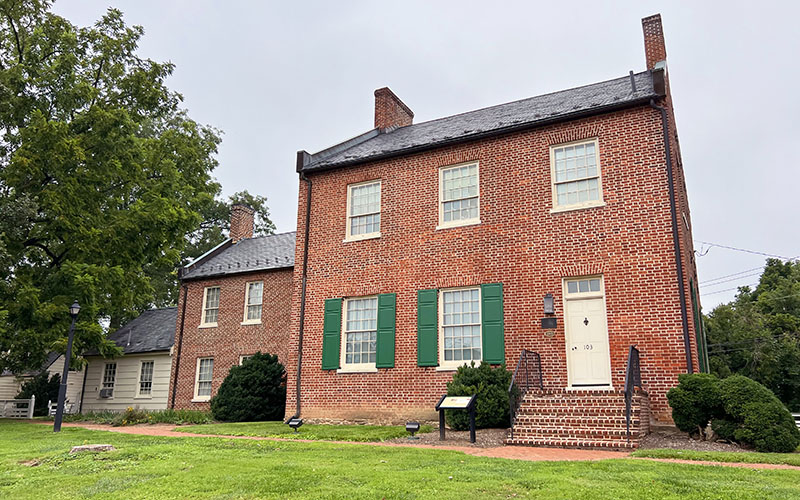 Two connected historic brick houses with green shutters and white trim sit on a grassy lawn with a few bushes and a tree nearby, under an overcast sky.