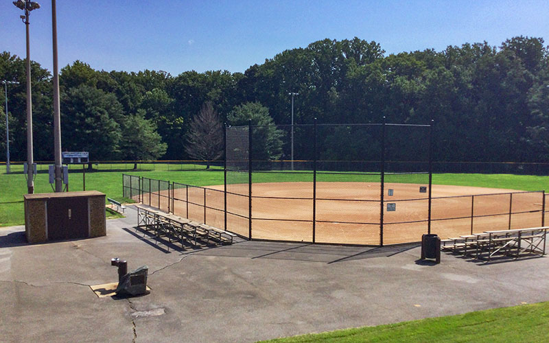 A baseball field with metal bleachers, a fenced backstop, scoreboard, and a small brick building, surrounded by green grass and trees under a clear sky.