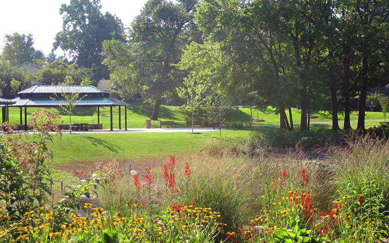 A park scene with a pavilion, picnic tables, grassy lawns, trees, and colorful flowers in the foreground on a sunny day.