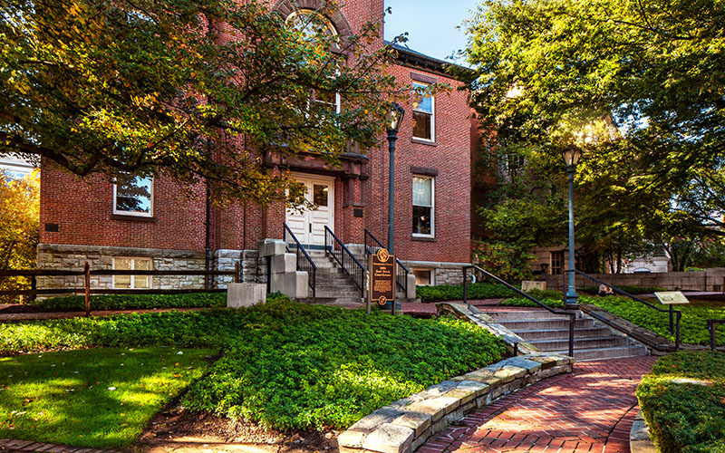 A brick academic building with a central white door, stone steps, and a sign, surrounded by trees, grass, and a brick walkway.