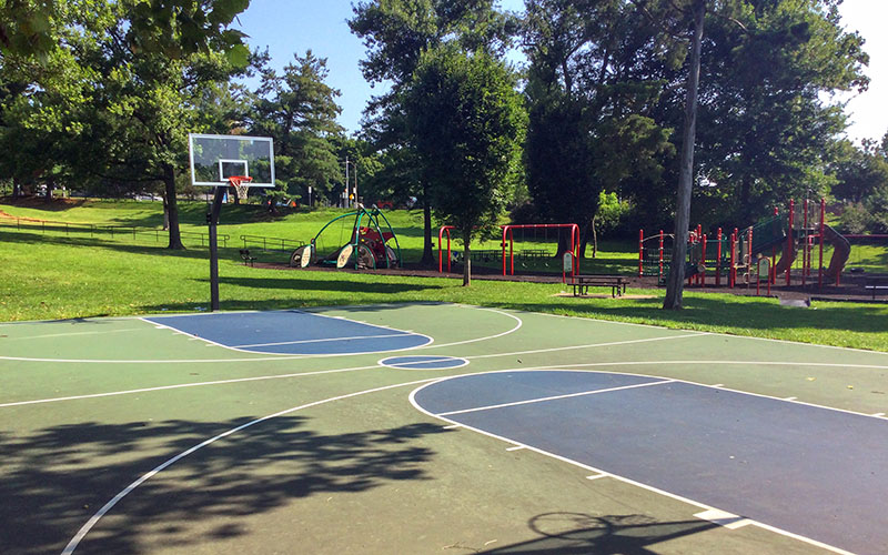 Outdoor basketball court with painted lines in a park, with playground equipment and trees in the background on a sunny day.