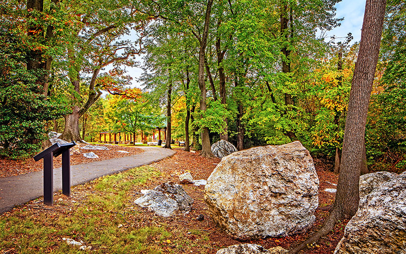A paved pathway curves through a wooded area with autumn foliage, large rocks, and an informational sign on the left.