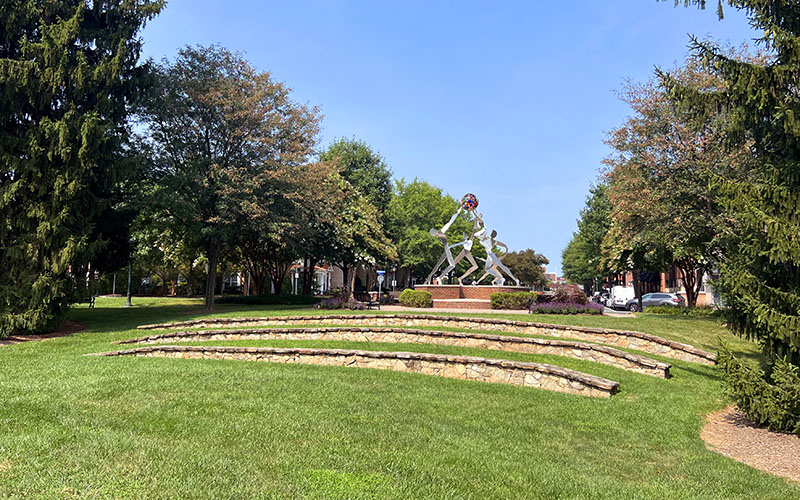 A tiered stone amphitheater faces a metal sculpture of human figures on a circular brick platform, surrounded by trees and green grass under a clear sky.
