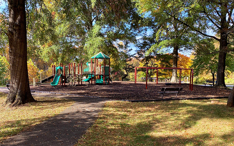 A playground with slides, climbing structures, and empty swings is surrounded by trees and shaded benches on a sunny day.