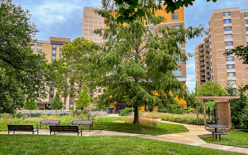 A landscaped urban park with benches and a pathway, surrounded by tall apartment buildings and trees under a partly cloudy sky.
