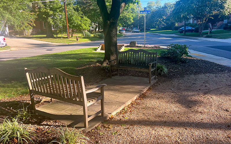 Two wooden benches face each other on a stone patio under a tree near a residential street intersection on a sunny day.