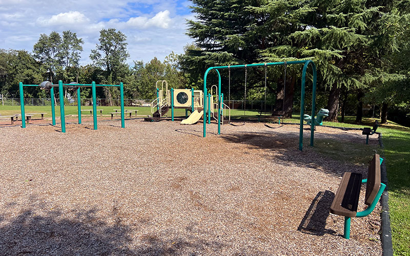 Empty playground with swings, a slide, climbing equipment, and benches, set on mulch ground with trees and grass surrounding the area.