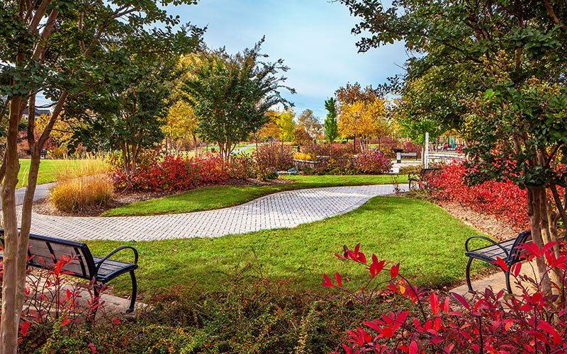Curved stone path runs through a landscaped park with benches, green grass, and trees displaying autumn foliage under a partly cloudy sky.