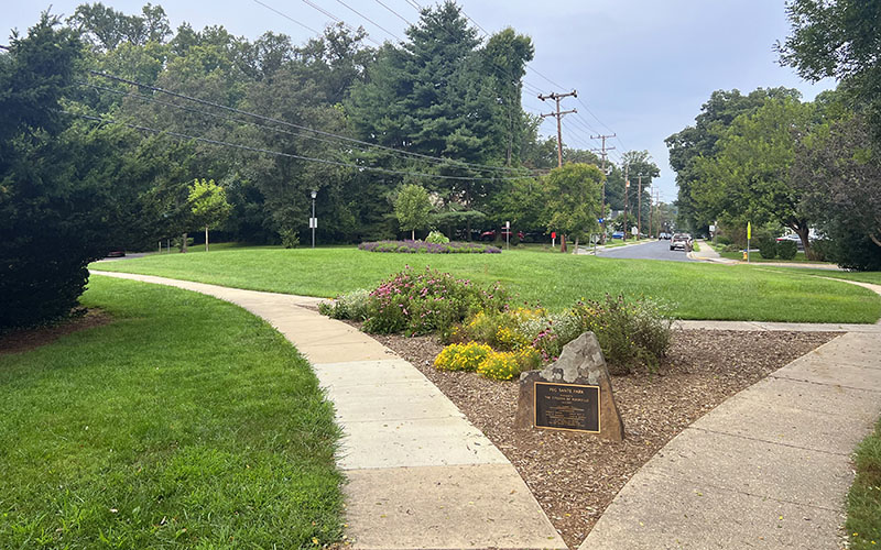 A small park with green grass, landscaped flower beds, intersecting sidewalks, trees, and a plaque mounted on a rock next to a main road with utility poles.
