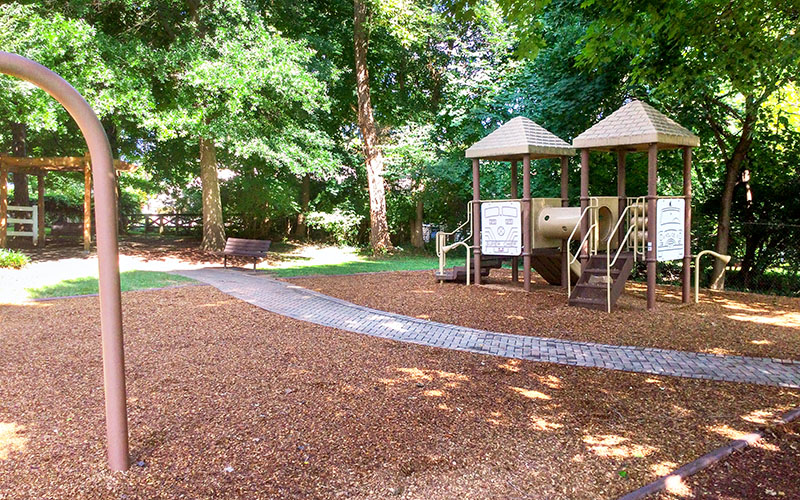 A playground with climbing structures, slides, a bench, and a shaded walking path surrounded by trees and wood chips on the ground.