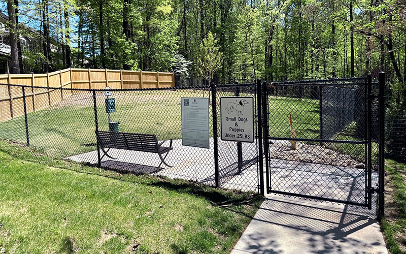 Fenced dog park area with a bench, waste station, and sign indicating it is for small dogs and puppies under 25 lbs, surrounded by trees.