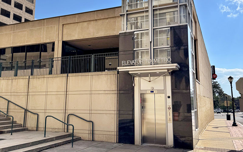 Street-level view of a modern elevator entrance labeled "Elevator to Metro" next to a beige building, with stairs and a railing on the left.