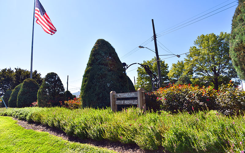 A manicured park with trimmed bushes, an American flag on a pole, and a wooden sign reading "Veterans Park City of Rockville" on a sunny day.