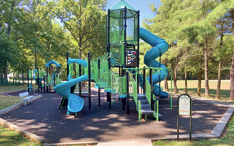 A modern playground with green and blue climbing structures, slides, and a spiral tube slide, surrounded by trees and benches on a sunny day.