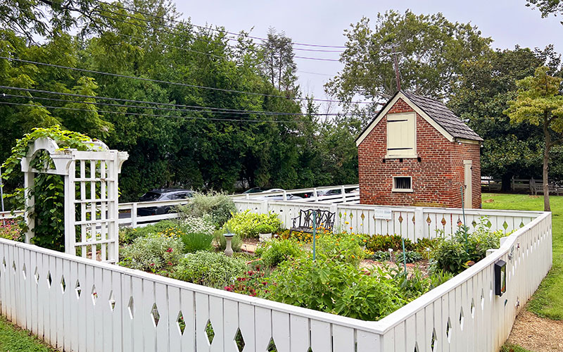 A white picket fence surrounds a well-kept garden with various plants and flowers; a small brick shed and arbor are inside the enclosure, with trees and a road in the background.