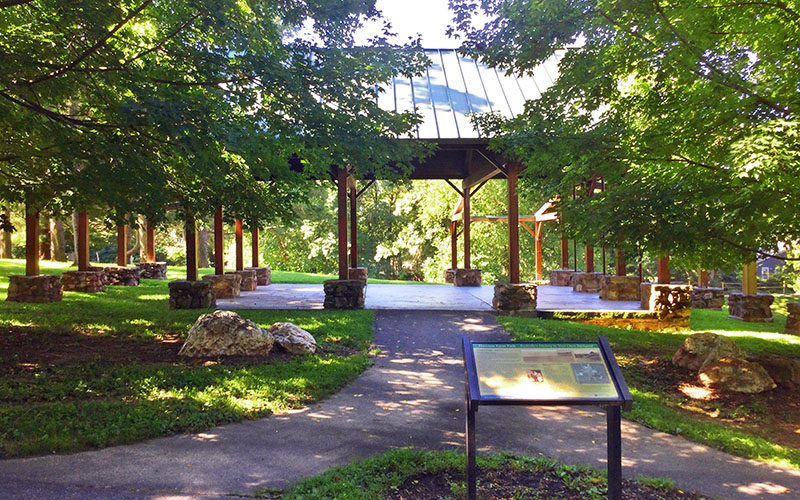 A shaded pavilion with a metal roof stands in a green park, surrounded by trees, stone posts, and a paved pathway with an informational sign in the foreground.