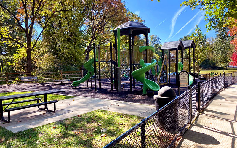 Fenced playground with green slides, climbing structures, and a picnic table, surrounded by trees on a sunny day.