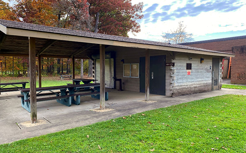 Covered picnic shelter with two tables next to a small building with restrooms, set on grass near trees with autumn foliage.