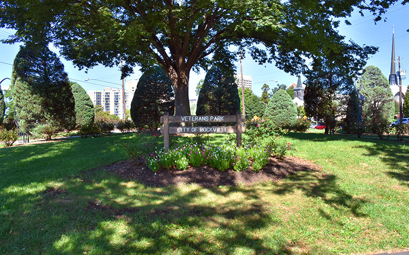 A wooden sign reading "Veterans Park, City of Rockville" stands in a landscaped area with flowers and bushes, surrounded by trees and buildings in the background.