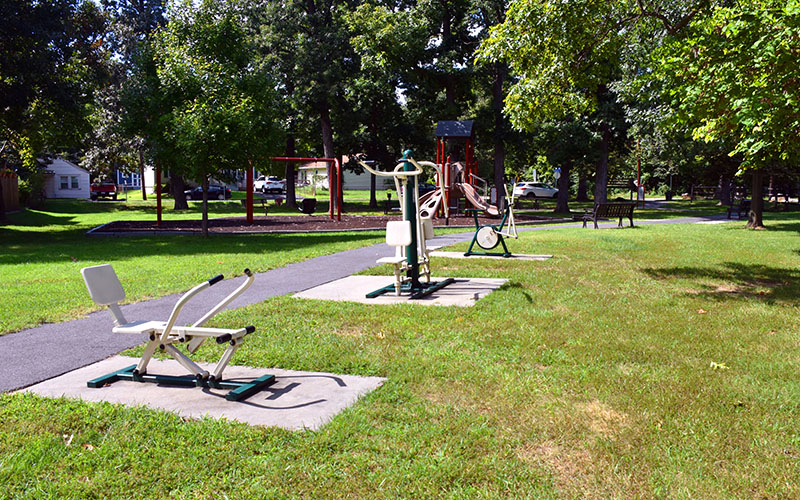 Outdoor fitness equipment is installed along a paved path in a grassy park, with trees and playground equipment visible in the background.