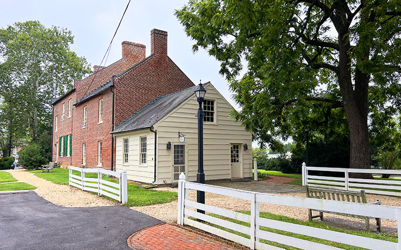 A historic two-story brick and wood house with green shutters, white fences, a bench, and large trees in a landscaped yard.