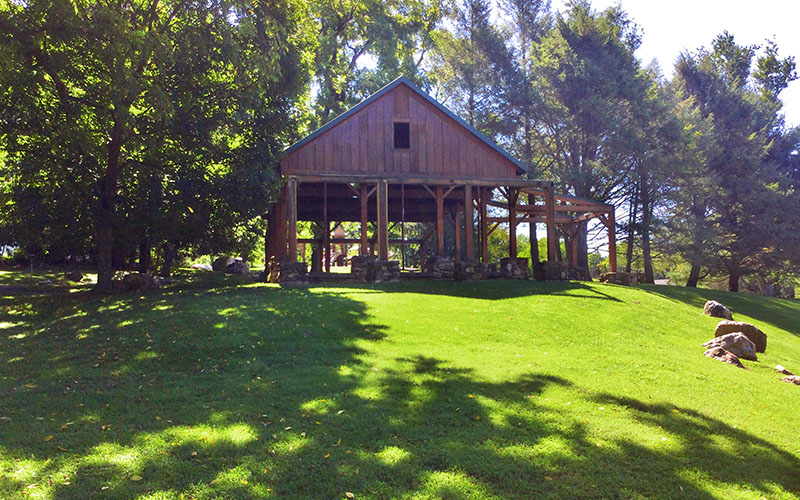 A wooden pavilion with open sides stands on a grassy lawn surrounded by trees, with sunlight filtering through the foliage.