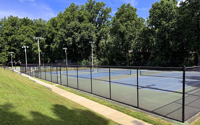 A fenced outdoor tennis court surrounded by trees and grass, with lights overhead and a clear blue sky above.