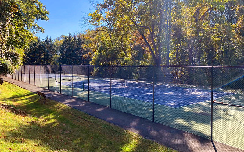 A fenced outdoor tennis court is surrounded by trees and grass on a sunny day.