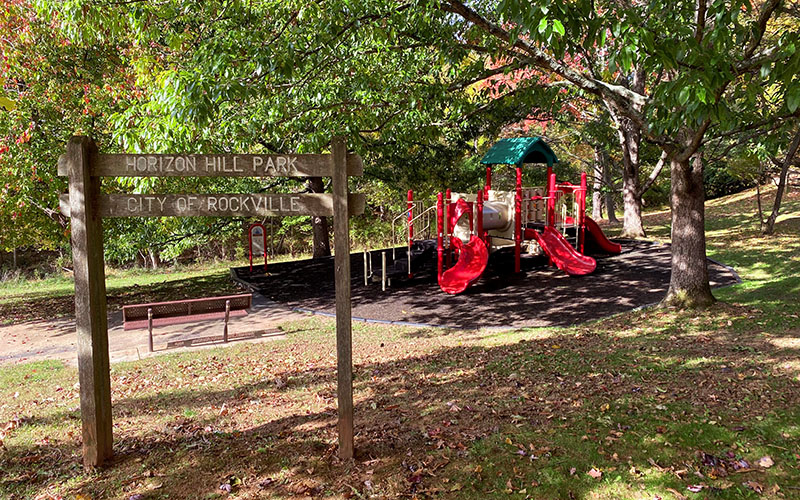 A wooden sign for Horizon Hill Park stands near a playground with red slides and a green canopy, surrounded by trees and grass.
