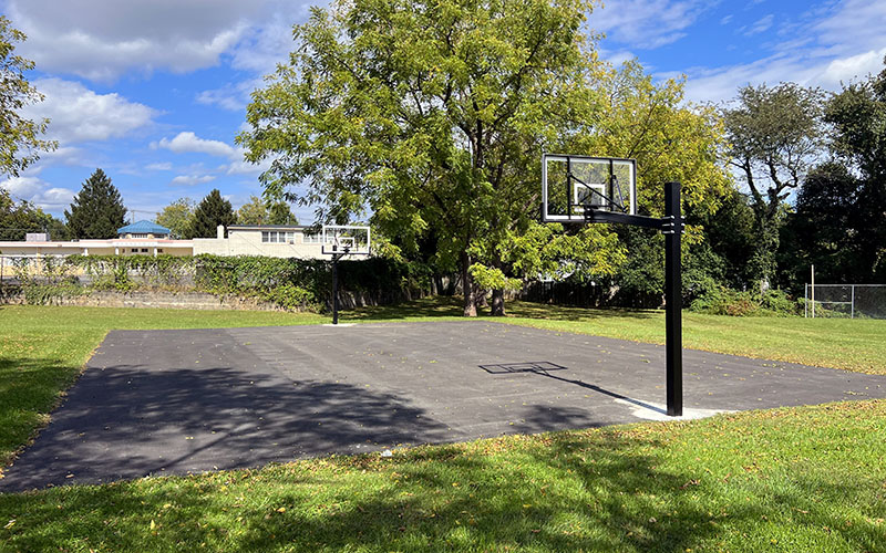Outdoor basketball court with two hoops, surrounded by grass and trees, under a partly cloudy sky. Some buildings are visible in the background.