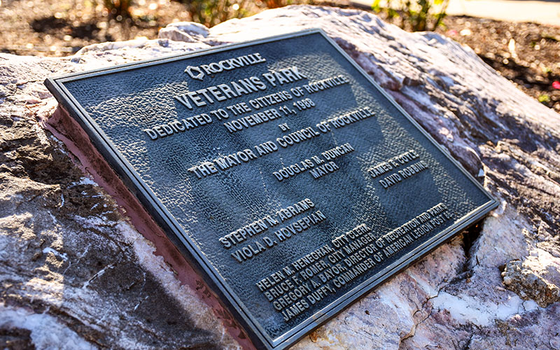 Bronze plaque on a large rock marks the dedication of Veterans Park in Rockville, honoring local officials and citizens, dated November 11, 1996.
