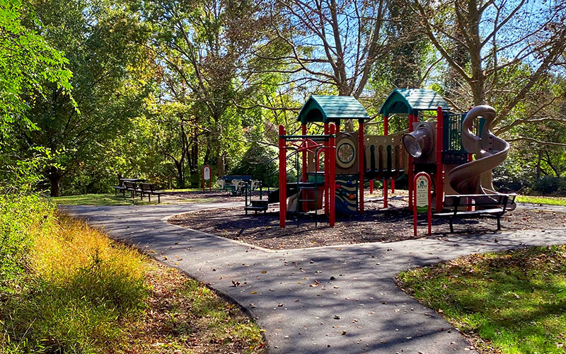A playground with slides and climbing structures sits next to a paved path, surrounded by trees and benches in a park setting.