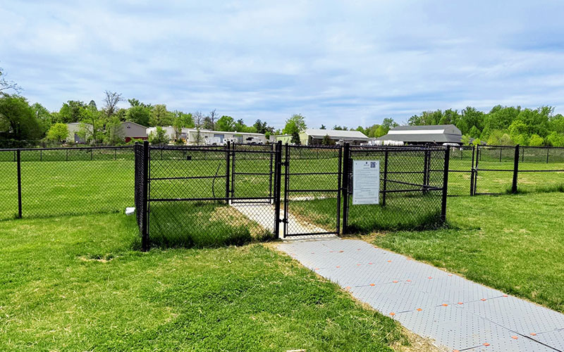 A fenced dog park entrance with a gate, sign, and paved walkway, surrounded by green grass and trees, with houses in the background under a partly cloudy sky.
