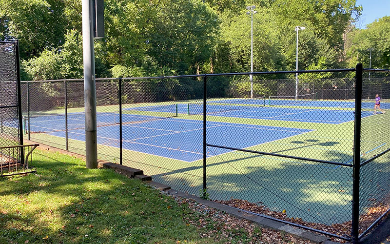Two blue and green outdoor tennis courts are enclosed by a black chain-link fence with trees and greenery surrounding the area. A person stands on the far court.