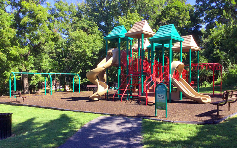 Outdoor playground with slides, swings, and climbing structures surrounded by trees and grass; benches and a trash can nearby under clear daylight.