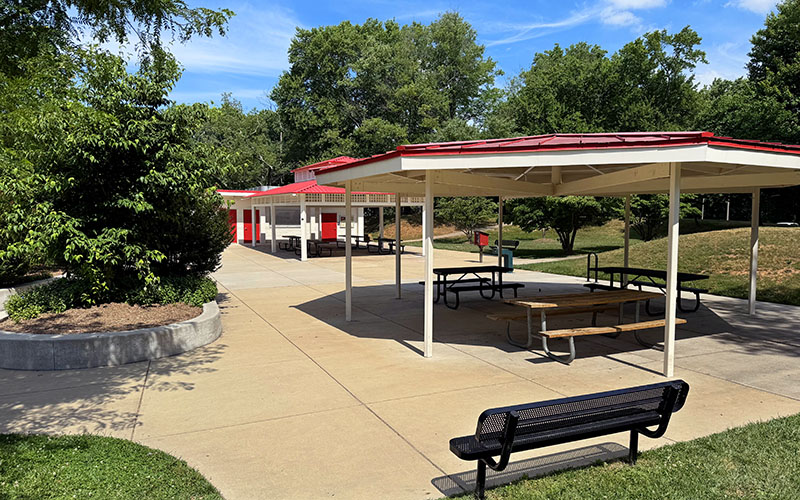 A shaded pavilion with picnic tables, a black bench, and a red-roofed building are surrounded by trees and greenery on a sunny day.