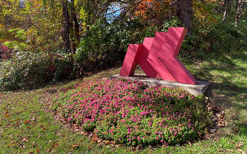 A red abstract metal sculpture stands on a concrete base next to a flower bed with pink flowers and surrounded by trees and greenery.