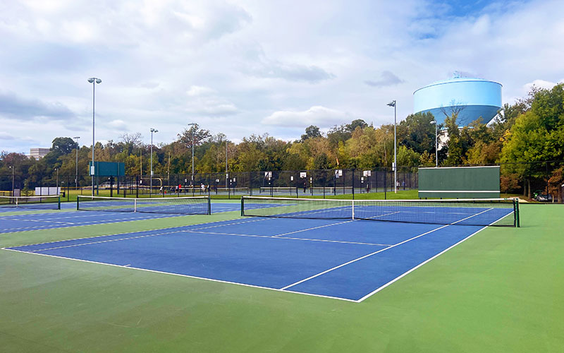 Outdoor tennis courts with blue and green surfaces, surrounded by fencing, trees, and a large blue water tower in the background under a partly cloudy sky.