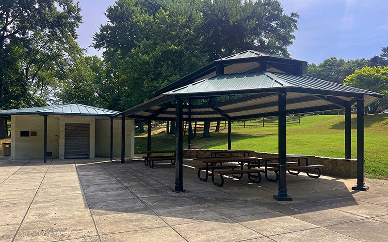 A covered pavilion with picnic tables stands next to a small building in a park with green grass and trees under a clear sky.