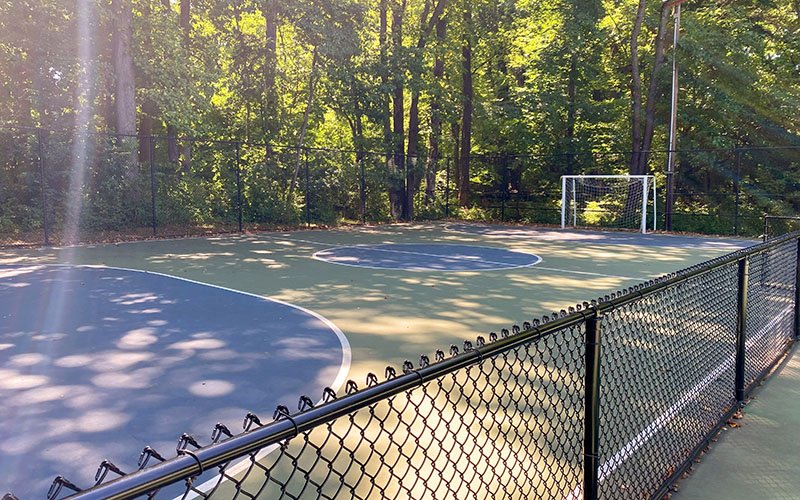 A fenced outdoor soccer court with a goal is surrounded by trees on a sunny day. Sunlight filters through the leaves, casting shadows on the surface.