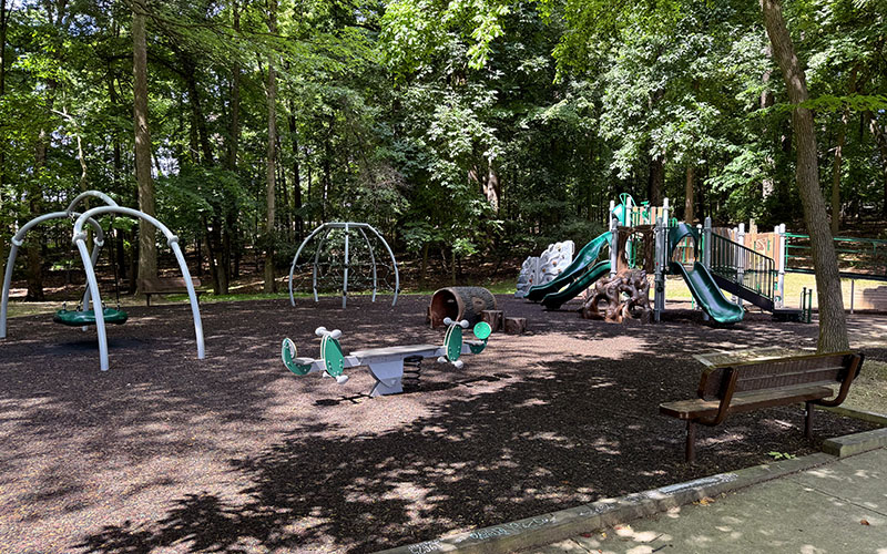 A playground with swings, climbing structures, slides, seesaws, benches, and trees providing shade in the background.