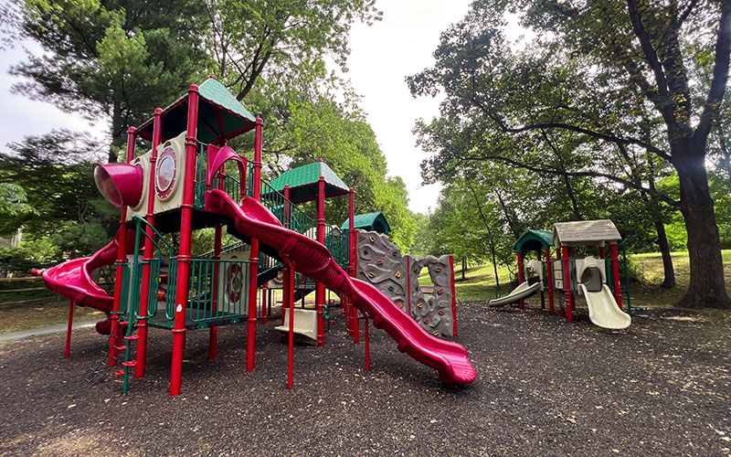 A playground with red and green play structures, including slides, climbing walls, and bridges, surrounded by trees and mulch ground covering.