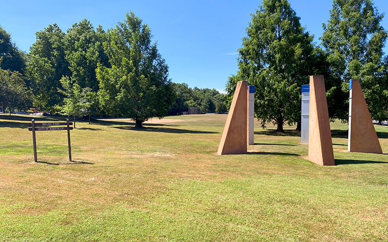 A grassy park with several trees, a brown wooden sign, and four large triangular stone structures on a sunny day.
