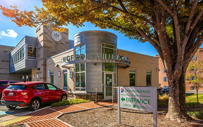 Modern building with large windows and "Rockville Police" sign; a red car is parked outside, and a sign directs visitors to the entrance and parking area.