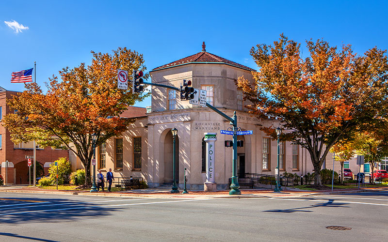 A small police station building with autumn trees in front, located at a street intersection with visible traffic lights and a U.S. flag in the background.