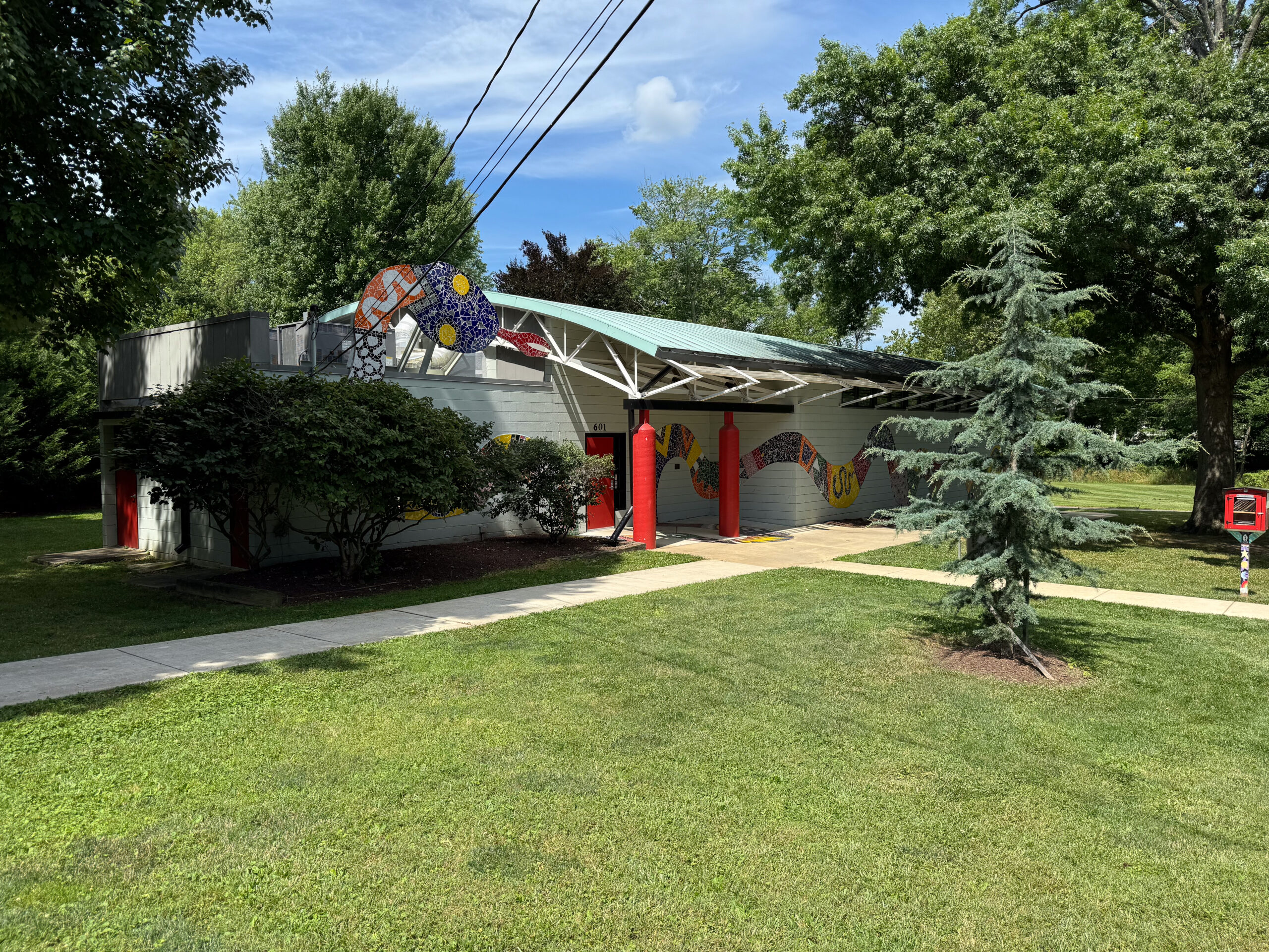 A modern building with colorful mosaic art on the exterior walls, surrounded by grass, trees, and a sidewalk under a partly cloudy sky.