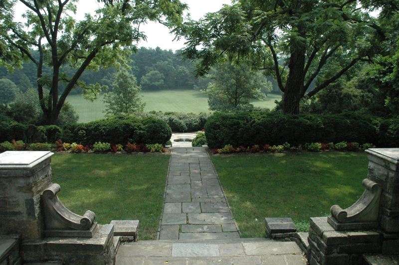 Stone steps lead down to a paved path, bordered by grass and shrubs, with trees framing a view of an open, grassy field in the background.